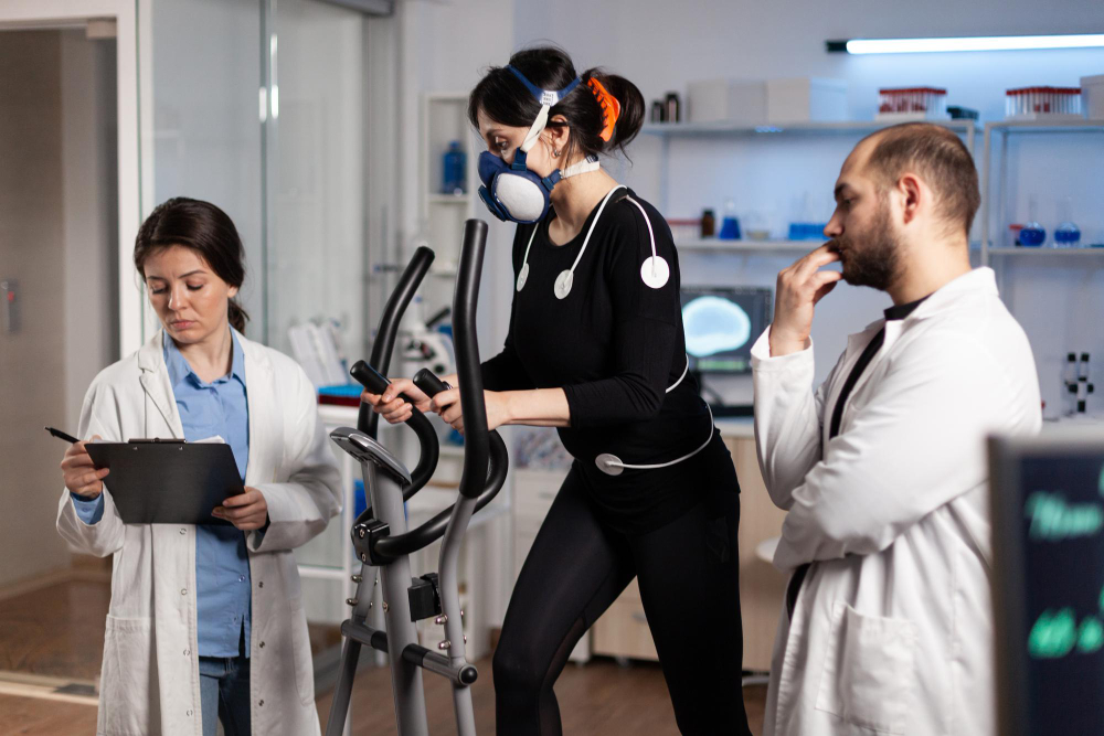 Une femme sur un vélo elliptique avec des électrodes et un masque de surveillance de la respiration sur elle, et avec deux chercheurs debout de chaque côté d'elle qui la surveillent.