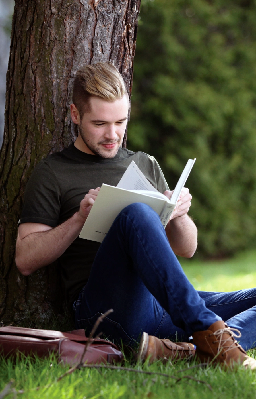 Un étudiant de l'Université Laurentienne assis à l'extérieur du campus, sous un arbre, en train de lire un livre.