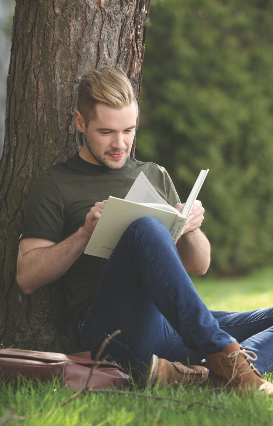 A Laurentian University student sitting outside on campus under a tree and reading a book.