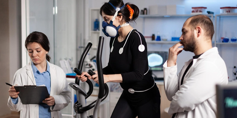 Une femme utilise un vélo elliptique équipé d'électrodes et d'un masque de surveillance respiratoire, tandis que deux chercheurs surveillent et collectent des données.