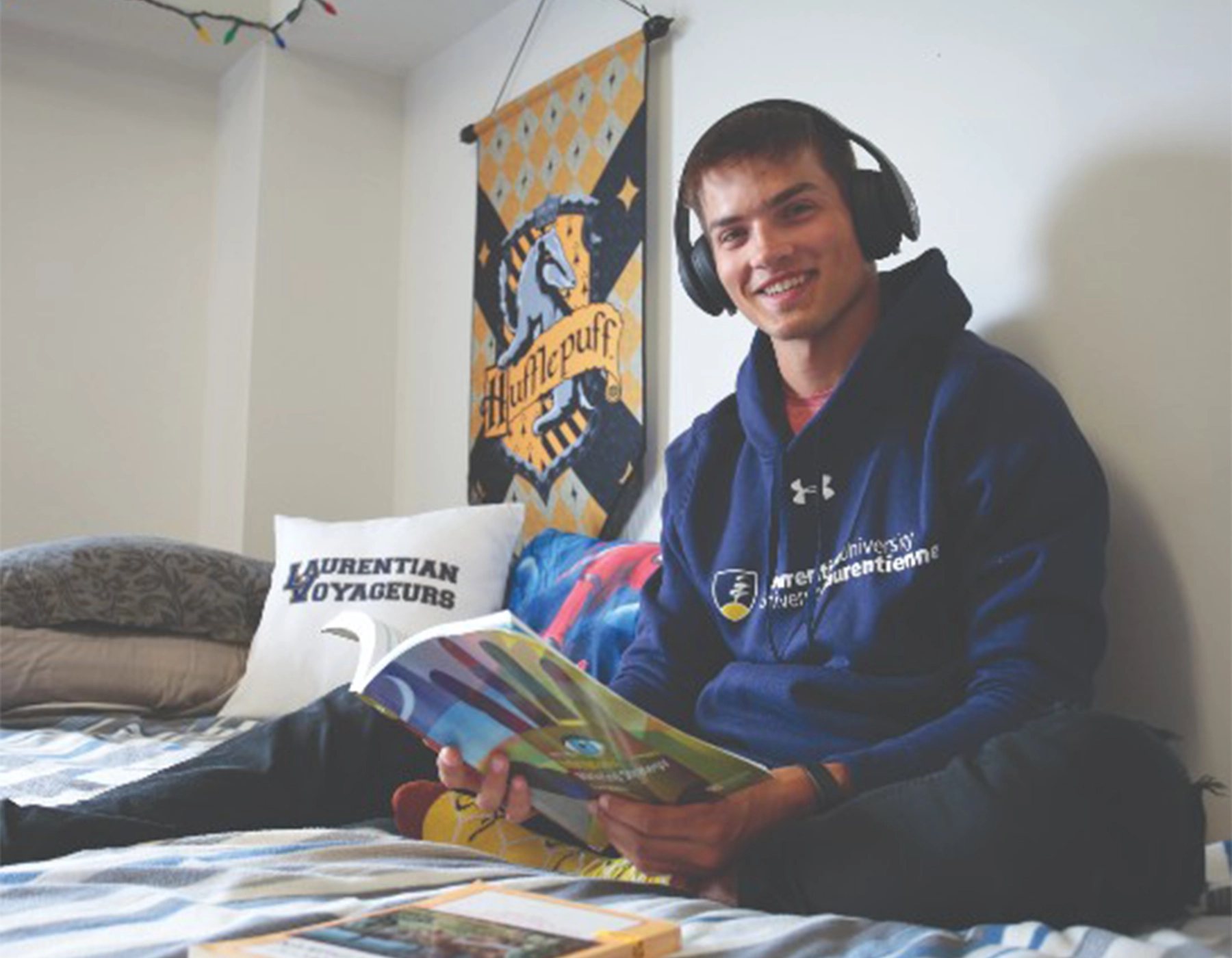 a student sits on his bed in residence. he is reading a book and has headphones on