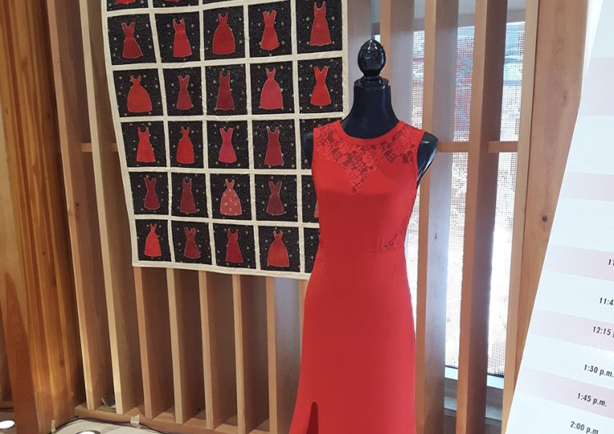 A long red dress displayed on a mannequin in the ISLC Round Room. On the wall behind it is a collage of photos of other red dresses.