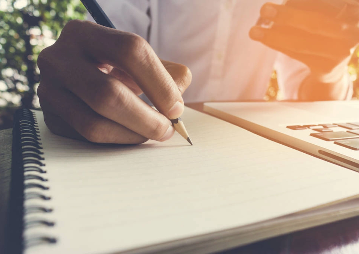 A hand holding a pencil, writing in a notebook.
