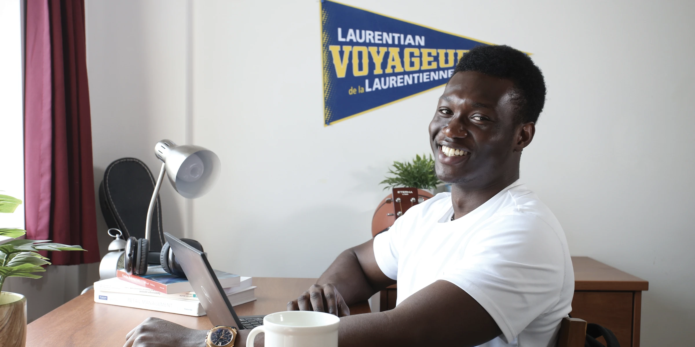 Un étudiant dans sa chambre en résidence universitaire, souriant alors qu'il travaille sur son ordinateur portable.