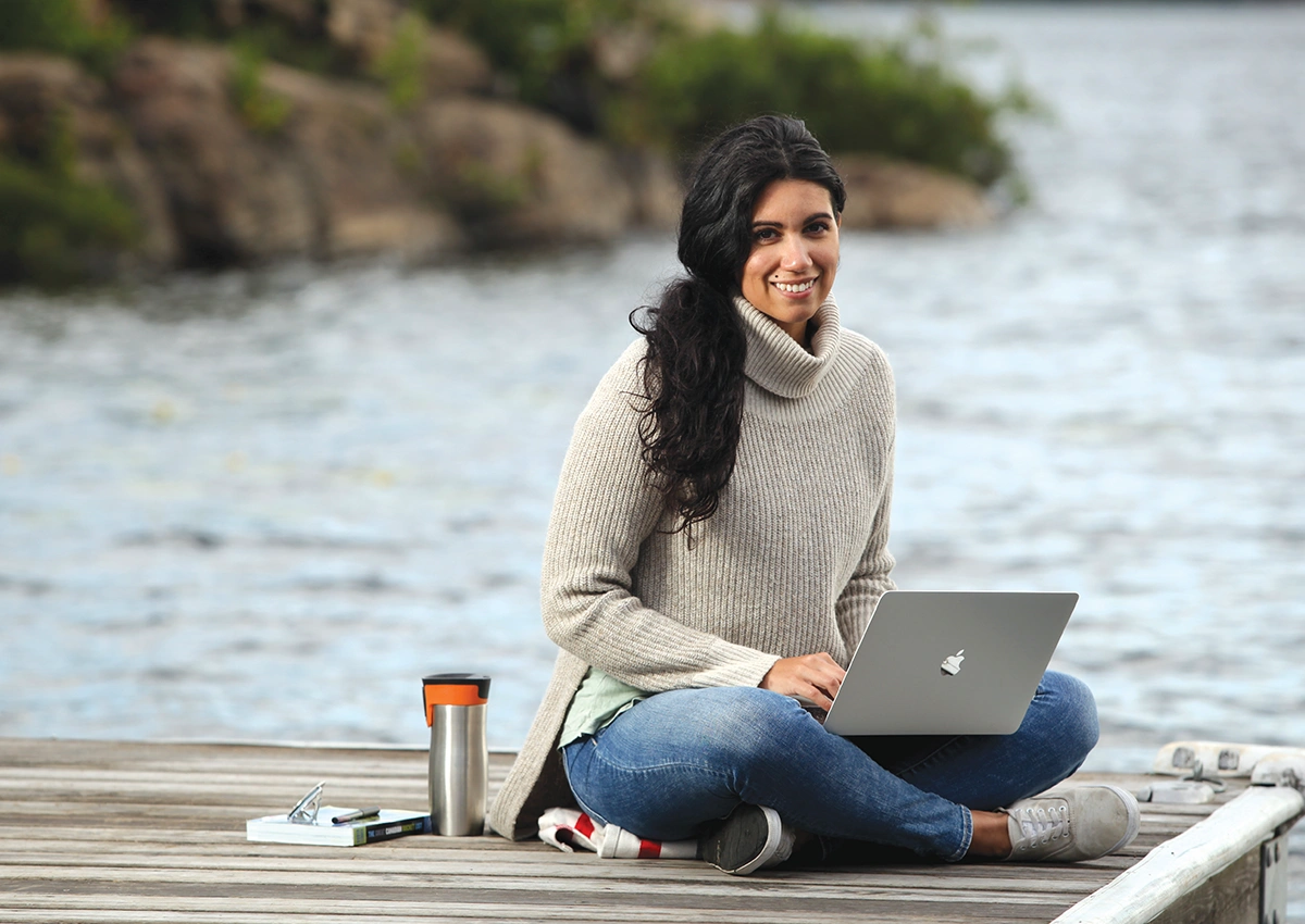 A Laurentian University student sitting on a dock by a lake while on her laptop.