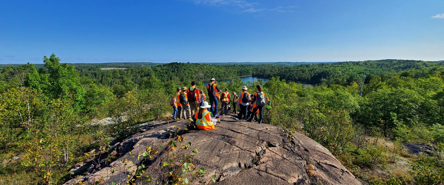 A group of students with a prof doing research on a large rock overlooking a forest.