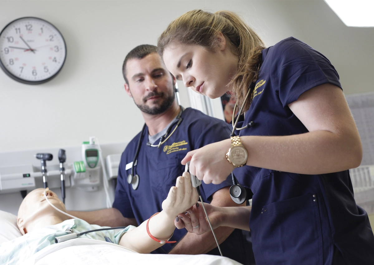 Nursing students applying a pulse monitor on the hand of a medical manikin.
