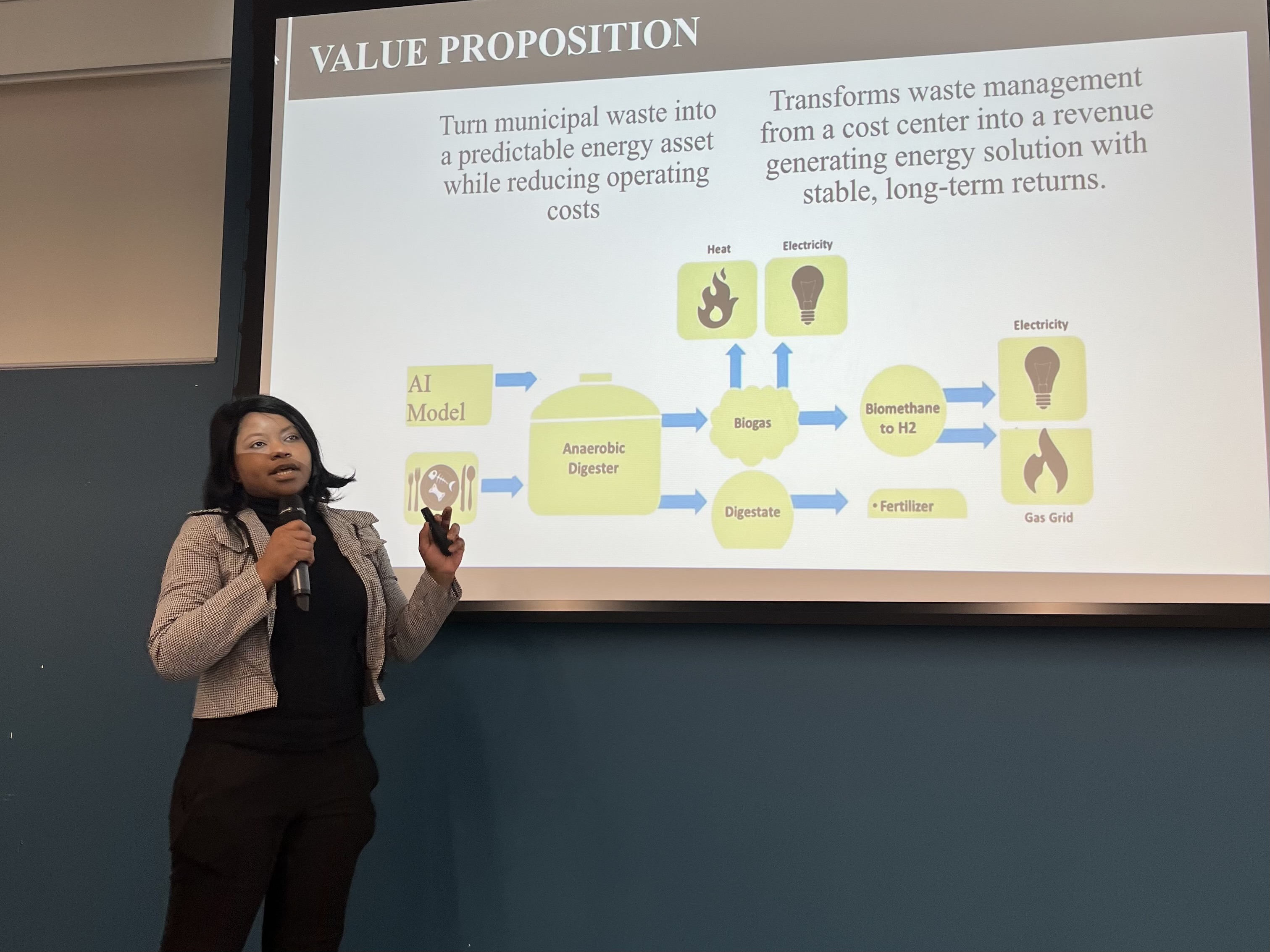 PhD student Jenny Alabi stands before a presentation slide titled "Value Proposition." She gestures toward a flowchart explaining how AI and anaerobic digestion transform municipal waste into renewable energy sources like heat, electricity, and fertilizer.
