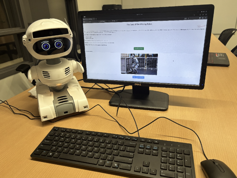 A wide shot of the small, white, humanoid Misty II robot on a wooden desk next to a computer monitor displaying a digital task. A black keyboard and mouse sit in the foreground.