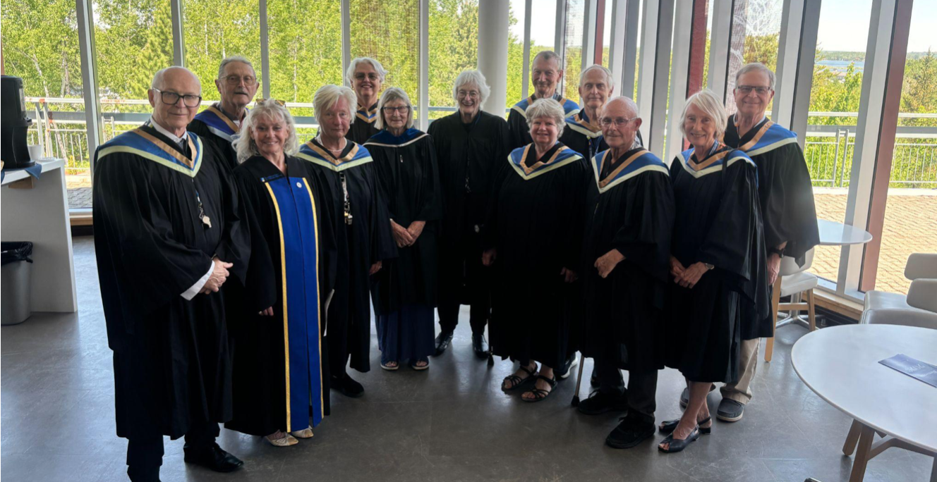 Alumni and retired faculty and staff wearing black and blue academic convocation gowns stand smiling together in a modern, bright building with large glass windows overlooking green trees. Dr. Pat Pickard stands in the center of the group.