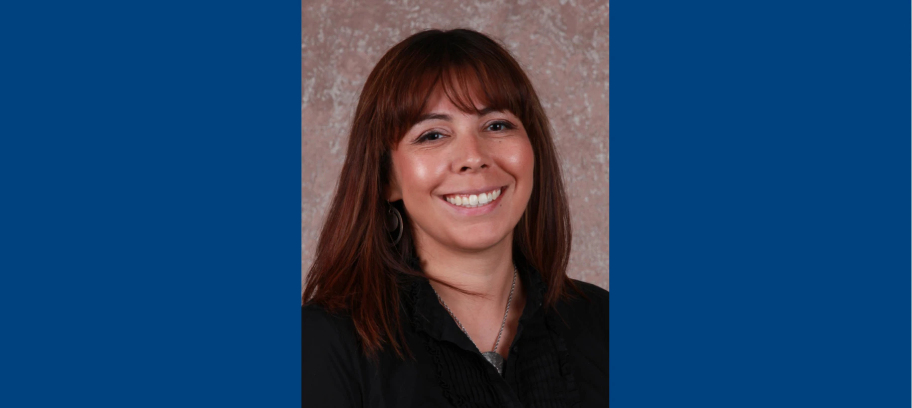 Headshot of Dr. Kona Williams smiling, featuring shoulder-length brown hair with bangs, a black collared shirt, and a silver necklace, set against a textured neutral background.
