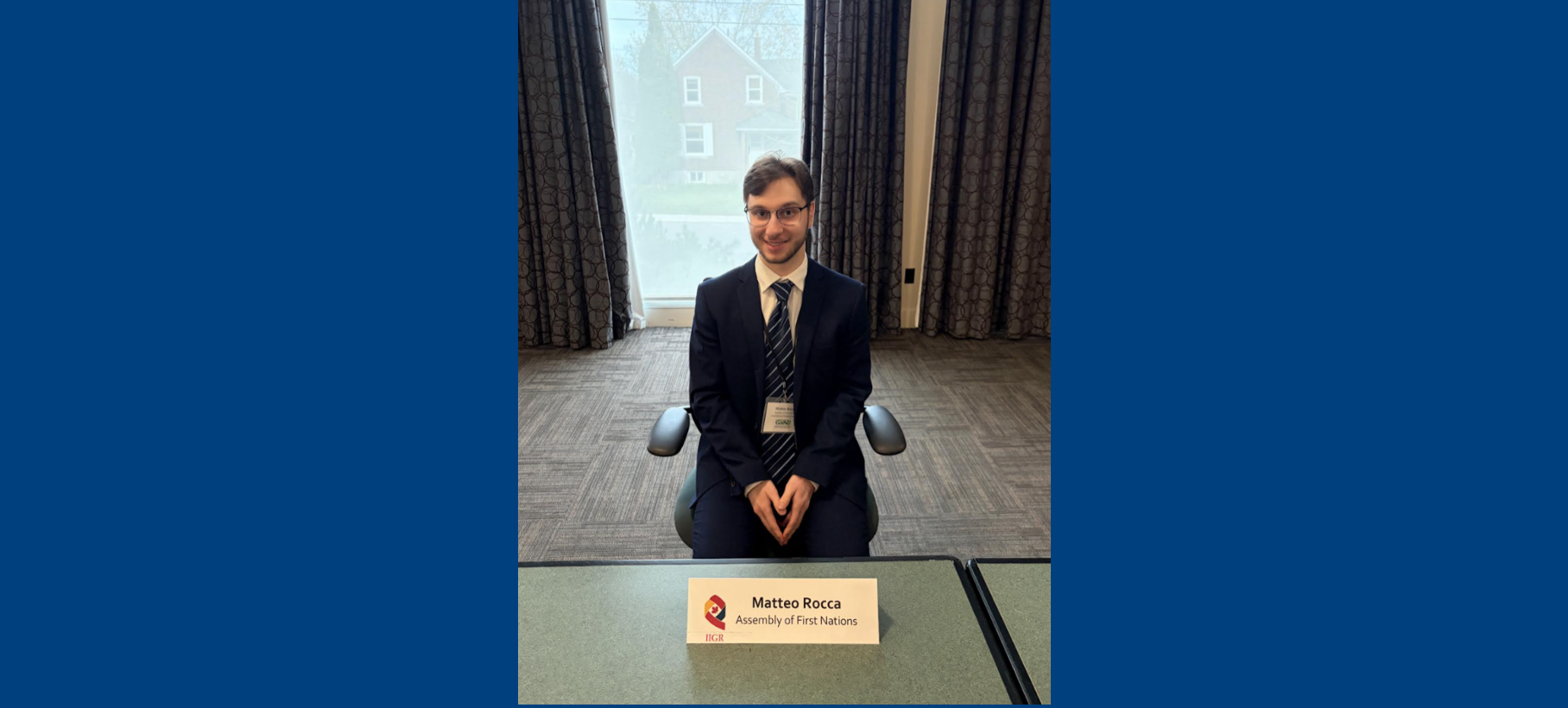 Laurentian University student Matteo Rocca sits smiling at a green conference table during the mock constitutional negotiations. He is wearing a navy blue suit jacket, white shirt, and striped tie. A printed table name card in front of him reads "Matteo Rocca, Assembly of First Nations.