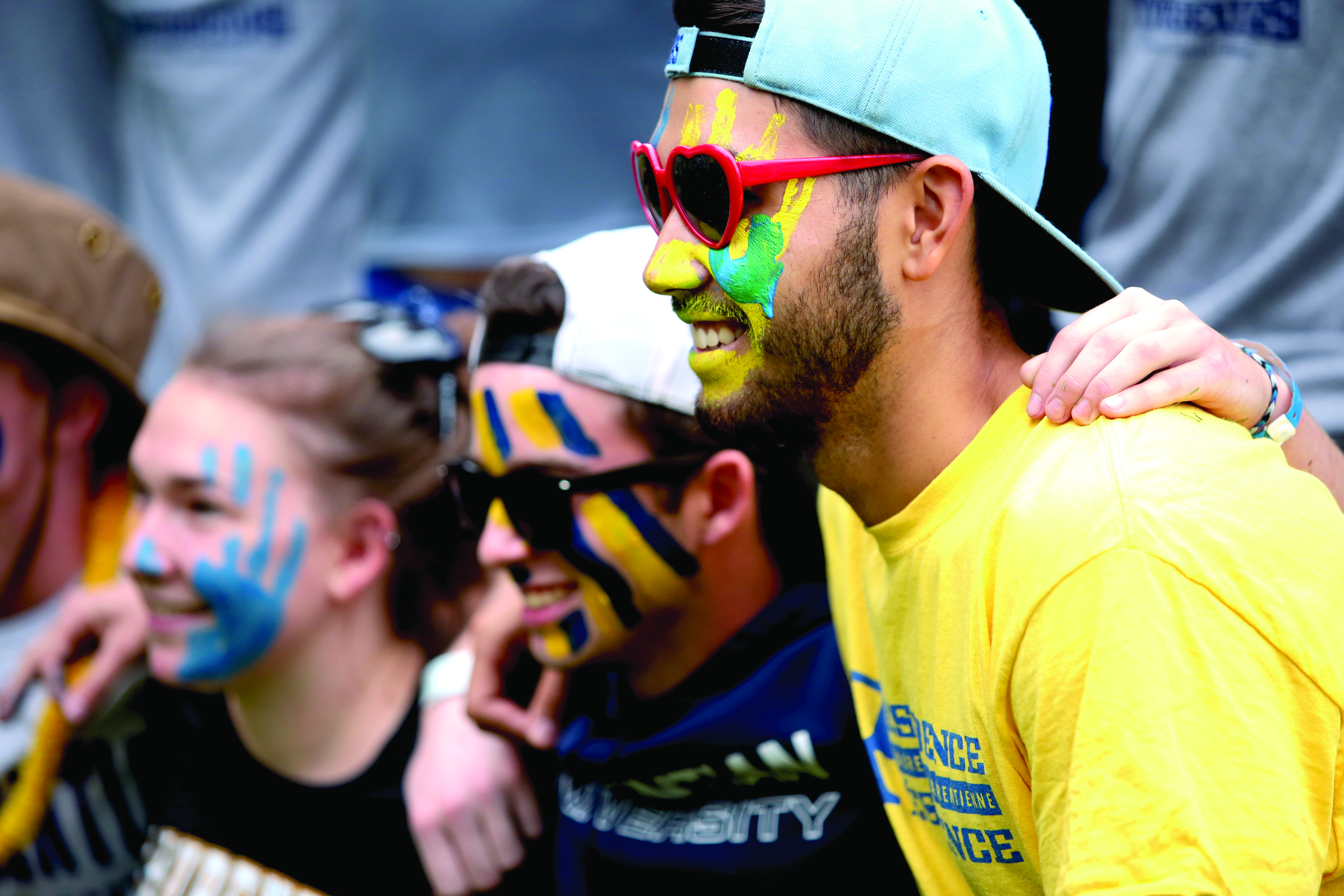 A close-up side profile of smiling Laurentian University students celebrating at an orientation event. The student in the foreground wears red heart-shaped sunglasses, a backwards blue baseball cap, a yellow t-shirt, and blue and yellow face paint, including a handprint painted on their cheek. Other students with blue and yellow face paint stand arm-in-arm beside them.