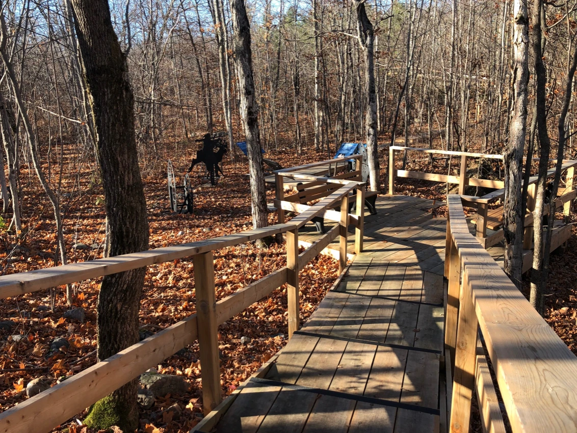 A wide view looking down a wooden boardwalk trail at the Sheguiandah Archaeological Site. The surrounding forest floor is blanketed with golden autumn leaves, and thin trees cast long shadows across the wooden path. Further down the trail, blue signage panels and dark metal silhouette sculptures are visible in the woods.