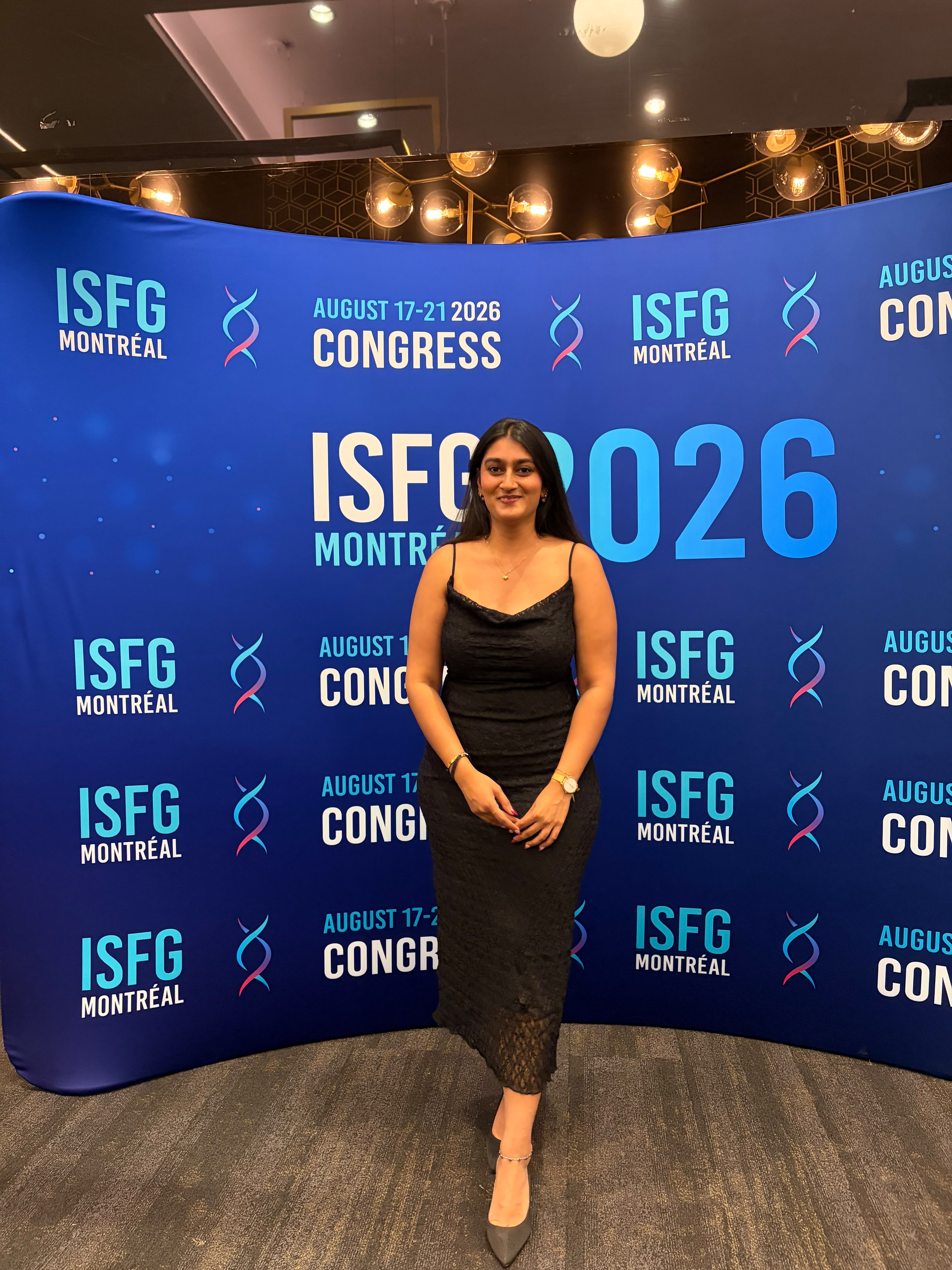 Shivani Gami posing in a formal black dress in front of a large blue backdrop repeating the "ISFG 2026 MONTRÉAL CONGRESS" logo with colorful DNA helix illustrations. 