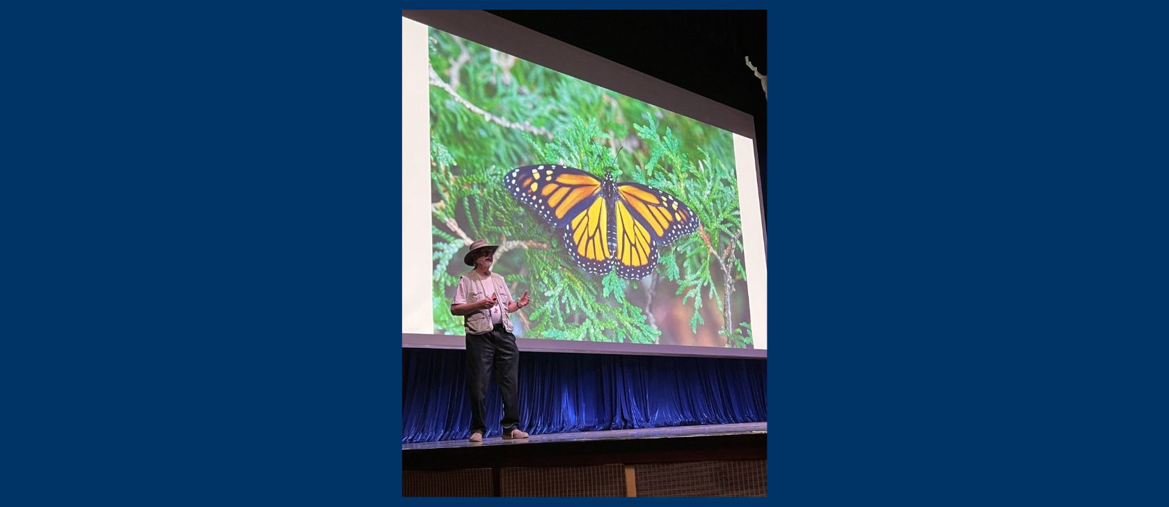 Dr. Joe Shorthouse presenting on stage in front of a large screen displaying a close-up photo of a monarch butterfly resting on cedar branches. He is wearing a wide-brimmed hat, beige vest, and dark trousers while gesturing to the audience.