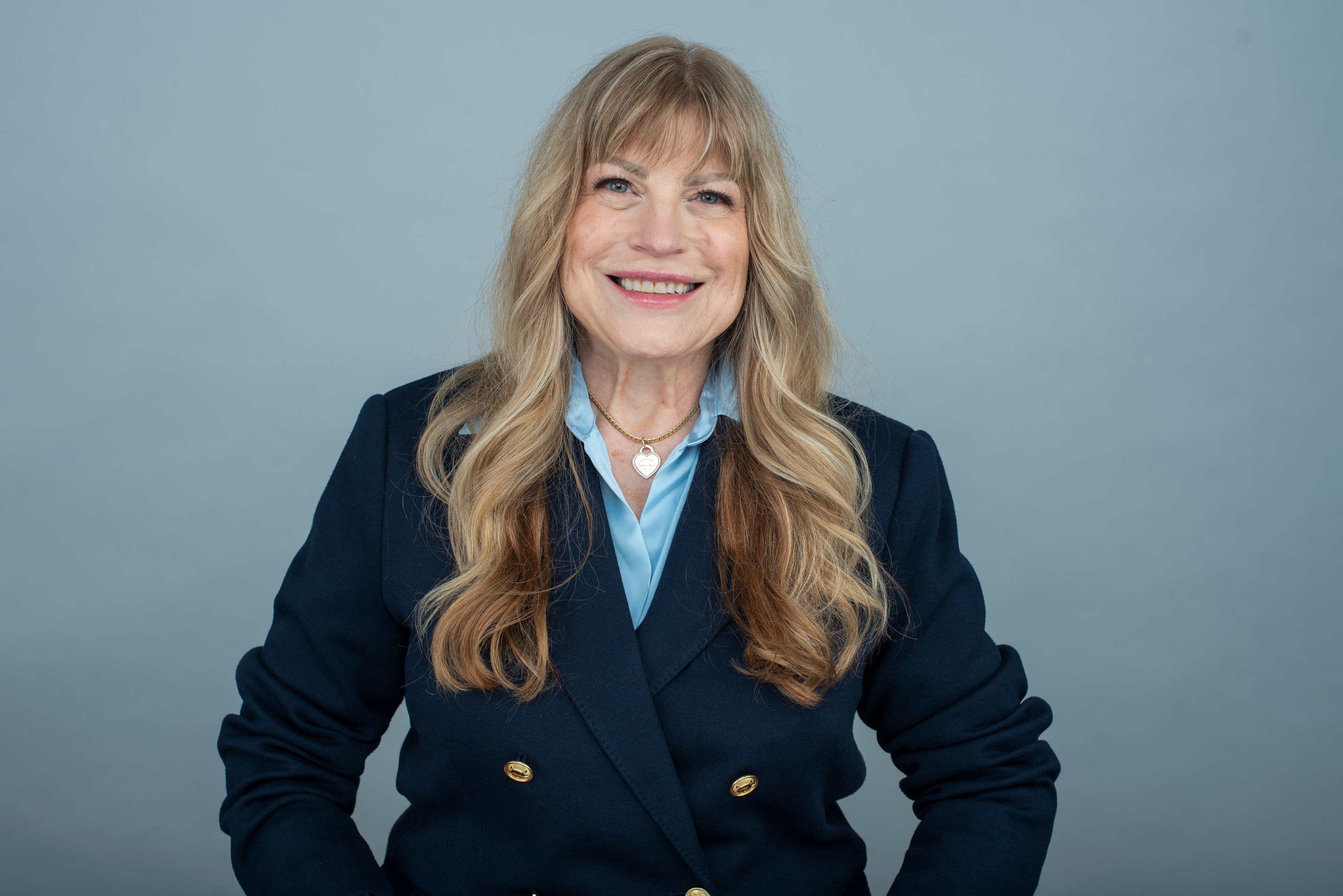 Professional studio portrait of Rachel Prudhomme smiling directly at the camera, wearing a dark navy double-breasted blazer with gold buttons, a light blue collared shirt, and long wavy blonde hair against a plain grey backdrop. 