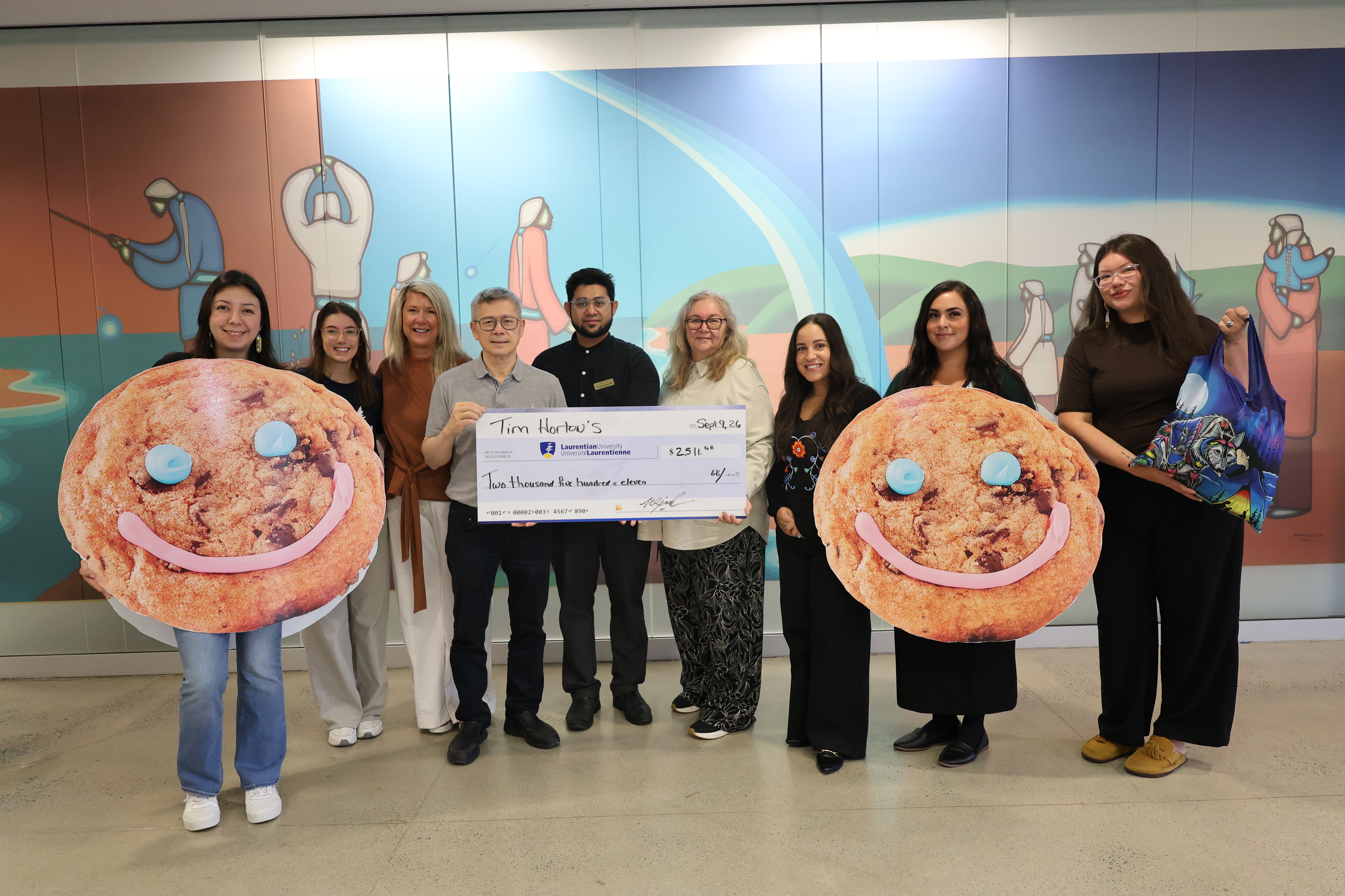 A group of nine people standing in front of an Indigenous wall mural, smiling and presenting a large ceremonial cheque for $2,511 to Laurentian University from Tim Hortons. Two individuals hold large cutouts of Tim Hortons Smile Cookies, while the person on the far right holds an example of a wellness kit.