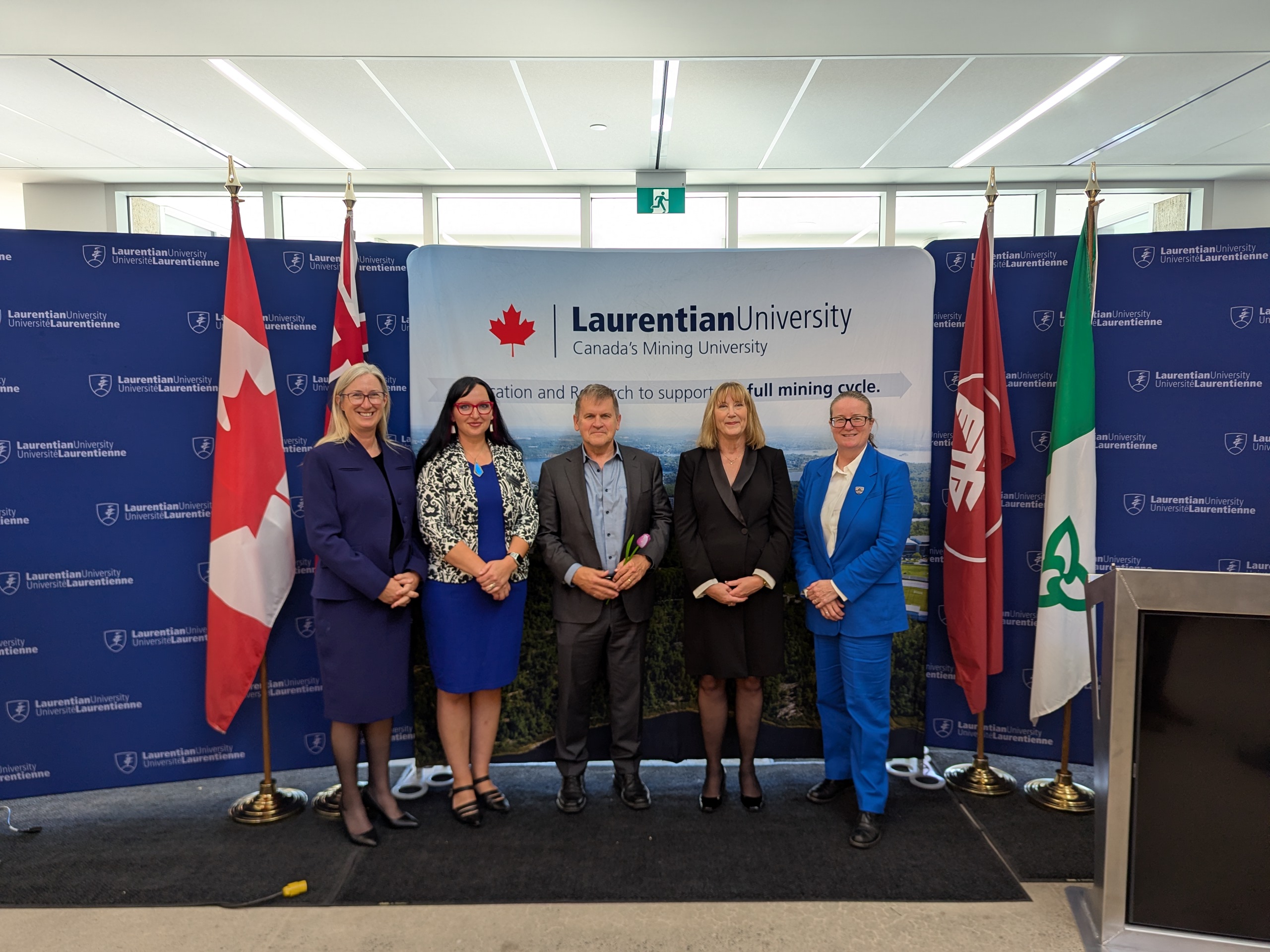 Five representatives pose in front of a Laurentian University Minerals and Mining Strategy backdrop, flanked by the Canadian, Ontario, Franco-Ontarian, and university flags.