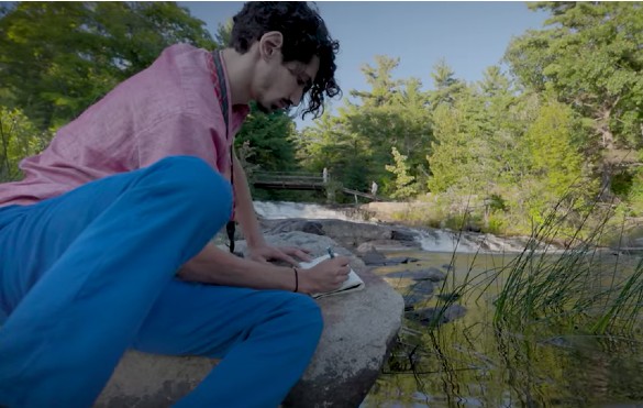 A student writing in a notepad on a rock near a river