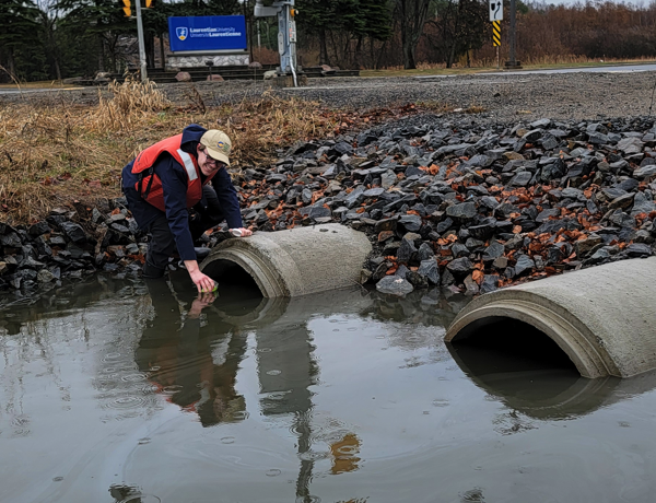Un chercheur portant un gilet de sauvetage prélève des échantillons d'eau des ponceaux devant l'enseigne de l'Université Laurentienne.