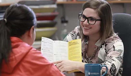 A woman smiling, showing a student an informative pamphlet.
