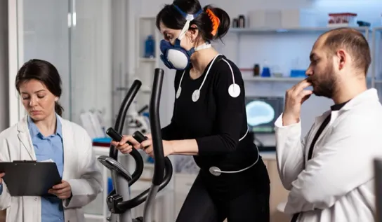 Une femme sur un vélo elliptique, équipée d'électrodes et d'un masque de surveillance respiratoire, avec deux chercheurs debout de chaque côté qui la surveillent.