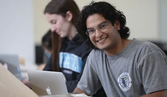 A student in lecture, smiling and taking notes on his laptop.