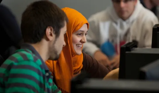 2 students sit in a classroom, looking at the same computer 