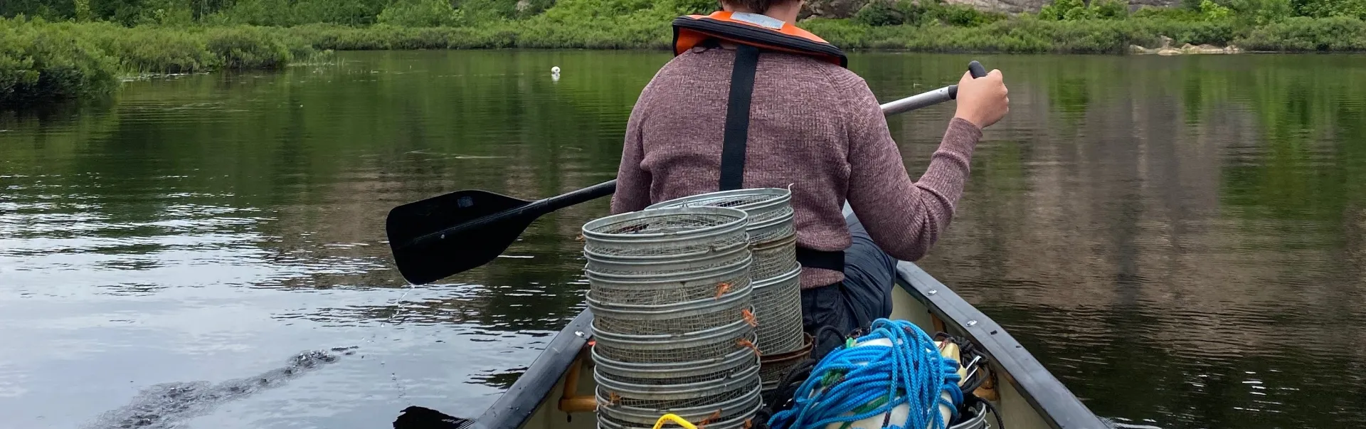 Person paddling a canoe filled with research gear on a calm river near rocky cliffs and forest.