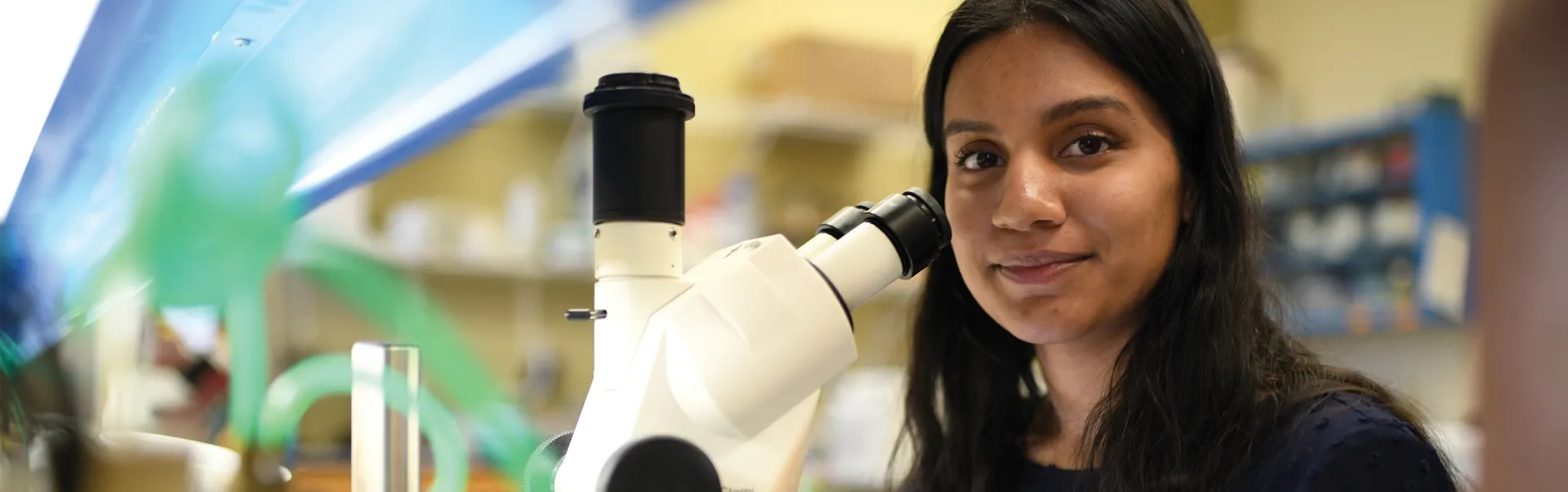 Student using a microscope, smiling at camera.