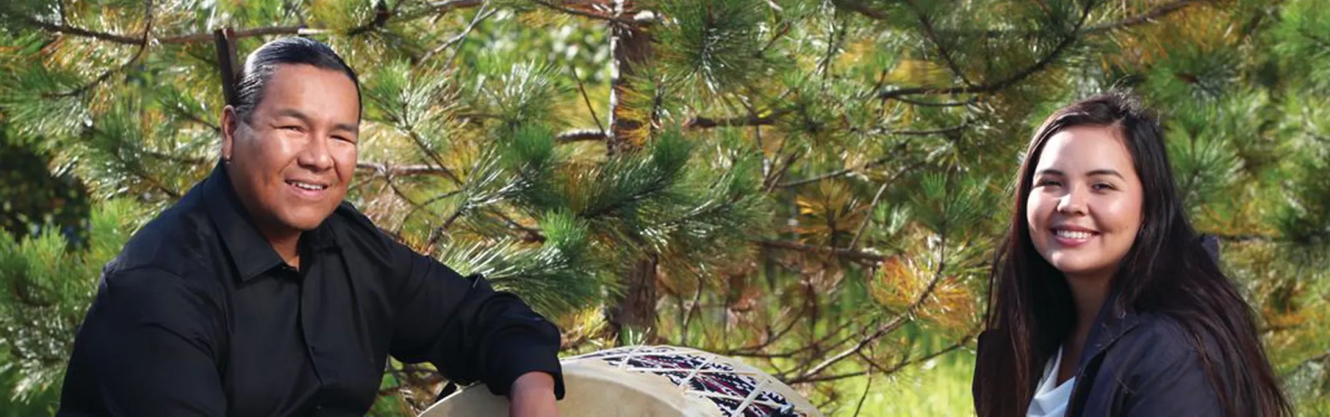 A man with a large, ceremonial drum sits across from a woman. A pine tree fills the background behind them.