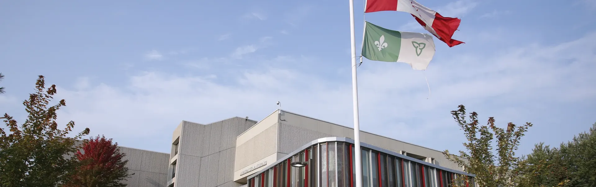 Édifice Alphonse-Raymond avec les drapeaux canadien et franco-ontarien flottant sur le mât à l'extérieur.