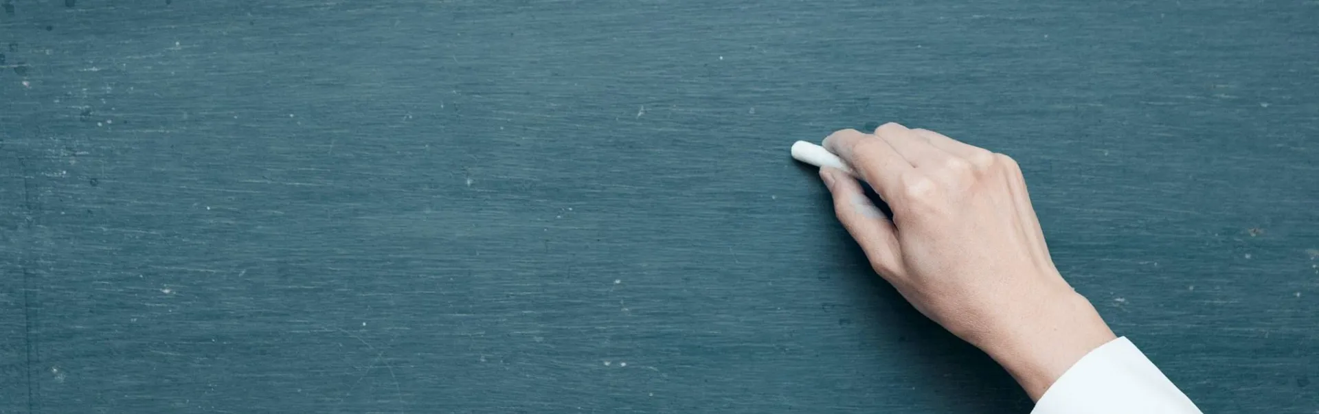 Hand holding white chalk poised at a green chalkboard