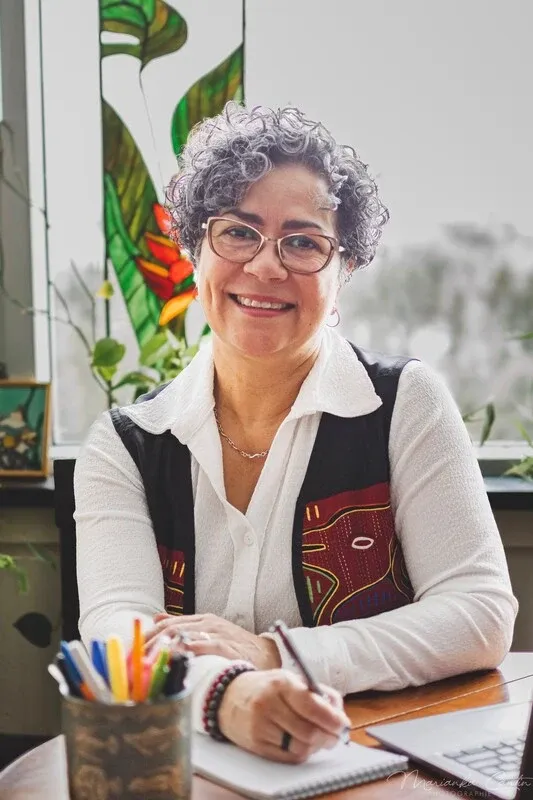A smiling person with short curly gray hair and glasses sits at a desk, writing in a notebook. They wear a white shirt and patterned vest, with a window and stained-glass decoration behind them.
