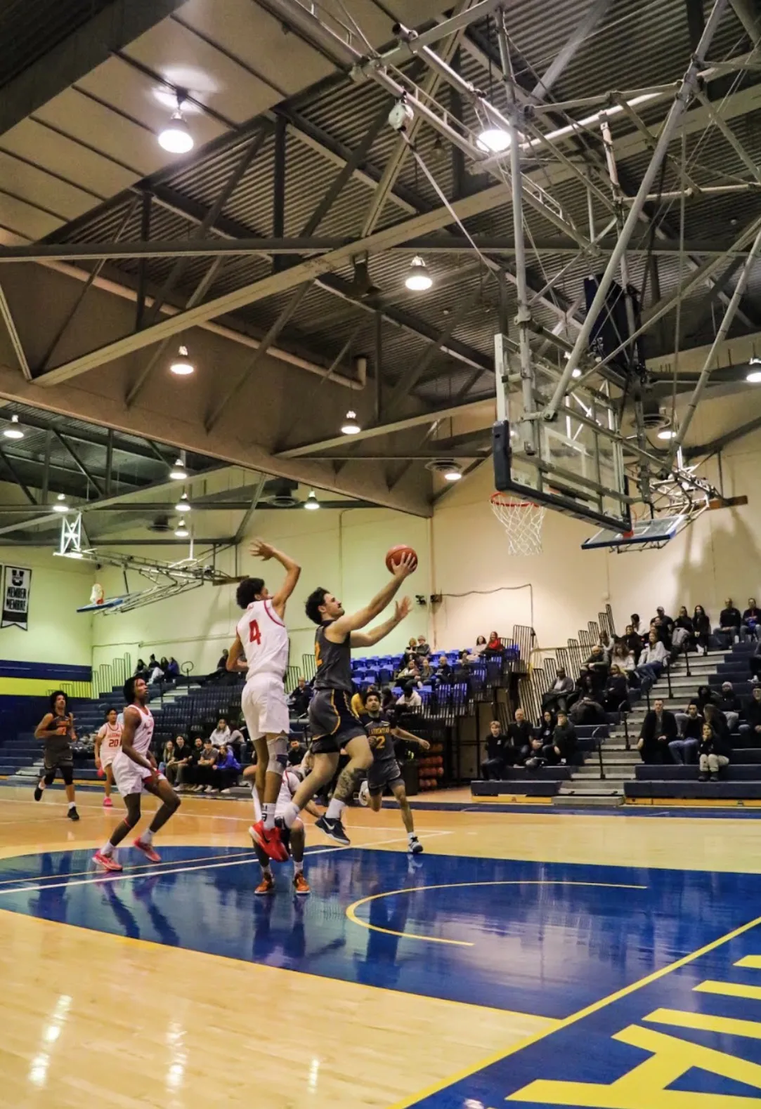An action shot from an indoor basketball game showing Evan Boudreau-Munro mid-air as he leaps toward the net to score a layup. He is wearing a dark grey Laurentian jersey and holding the basketball aloft with both hands, while an opposing player in a white jersey jumps beside him to block the shot. 