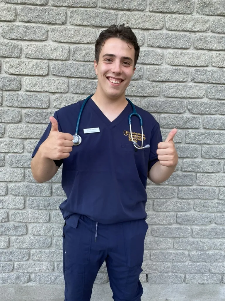 Jacob Sintic, posing in front of a grey stone brick wall. He is wearing dark blue medical scrubs and has a blue stethoscope draped around his neck. He is giving two thumbs up to the camera.