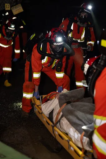 Laurentian University Mine Rescue team members perform a simulated underground casualty evacuation. The students wear orange high-visibility coveralls, breathing apparatuses, and helmets with lit lamps while securing a patient on a yellow stretcher.