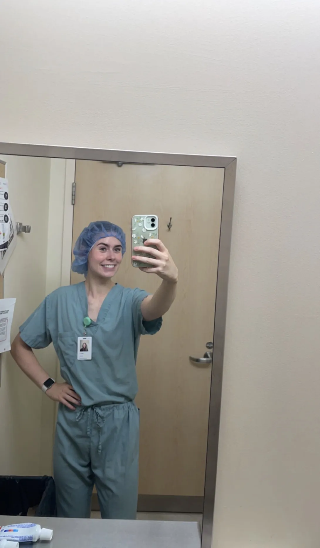 A mirror selfie of Alex Kowalski taking a photo on her smartphone during her hospital training. She is smiling and wearing light teal medical scrubs and a blue disposable surgical hair net.