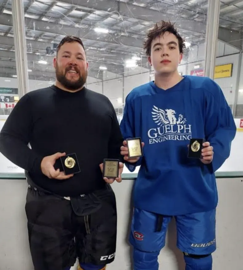 Christopher Clouthier standing alongside a younger teammate inside a hockey arena, both smiling and holding up award plaques and medals. Clouthier, on the left, has a dark beard and wears a black long-sleeved athletic top with hockey pants. The teammate on the right wears a blue hockey jersey with "Guelph Engineering" and a griffin crest printed on the front, along with blue hockey pants and gloves. ____________________________