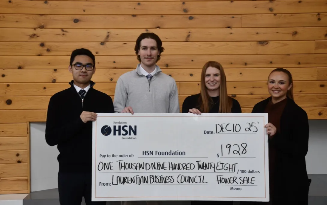 Four individuals stand in a line against a light-coloured wooden panelled wall, smiling while holding a large, oversized donation check from the Laurentian Business Council Flower Sale. Paul Petitclerc stands second from the left in a light grey quarter-zip sweater and tie. The check is made out to the HSN Foundation for the amount of $1,928, dated December 10, 2025.