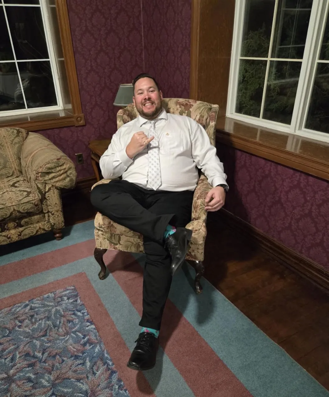 A relaxed indoor portrait of Christopher Clouthier sitting cross-legged in a floral-patterned armchair in a room with patterned magenta wallpaper and wood trim. He is smiling widely, wearing a white dress shirt, a light-coloured patterned tie, black dress pants, and patterned teal socks, while making a hand gesture toward the camera.