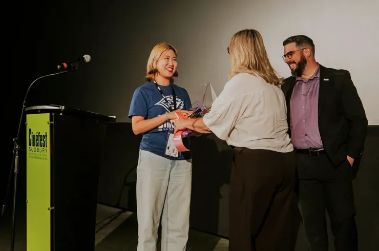 Felice Guo smiling on stage at a microphone podium during Cinefest Sudbury, wearing a blue volunteer t-shirt and acceptance lanyard. She is being presented with a bouquet of flowers by festival officials in front of a cinema screen, celebrating her recognition as the Youth Volunteer Award Winner. 