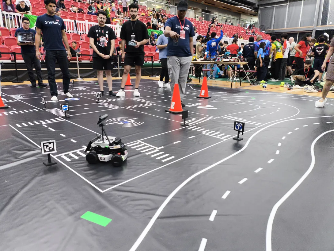 A small, four-wheeled autonomous vehicle labeled number 03 navigates a simulated road course map on a gymnasium floor during a competition. Members of the Laurentian SnoBots team stand in the background, carefully watching the vehicle's run.