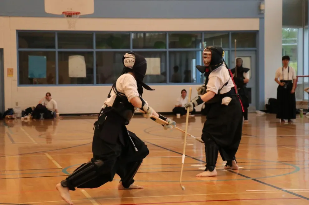 wo martial artists engaged in a dynamic practice session inside a gymnasium. They are wearing full traditional protective armour (bogu) and using long wooden weapons called Naginata, representing a practice session of the Naginata.