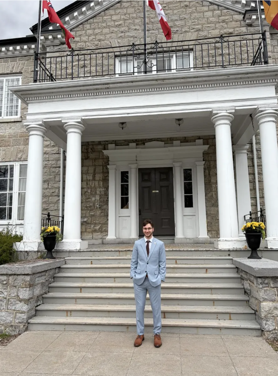 Full-length portrait of Matteo Rocca smiling while standing on the grand stone steps of a historic columned building. He is wearing a sharp, light blue suit with a white shirt, dark tie, and brown dress shoes.