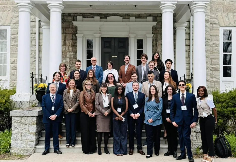 A group photo of twenty-two university student delegates and faculty advisors posing together on the wide stone steps under the large white columns of a historic estate building in Kingston during the national mock negotiations.