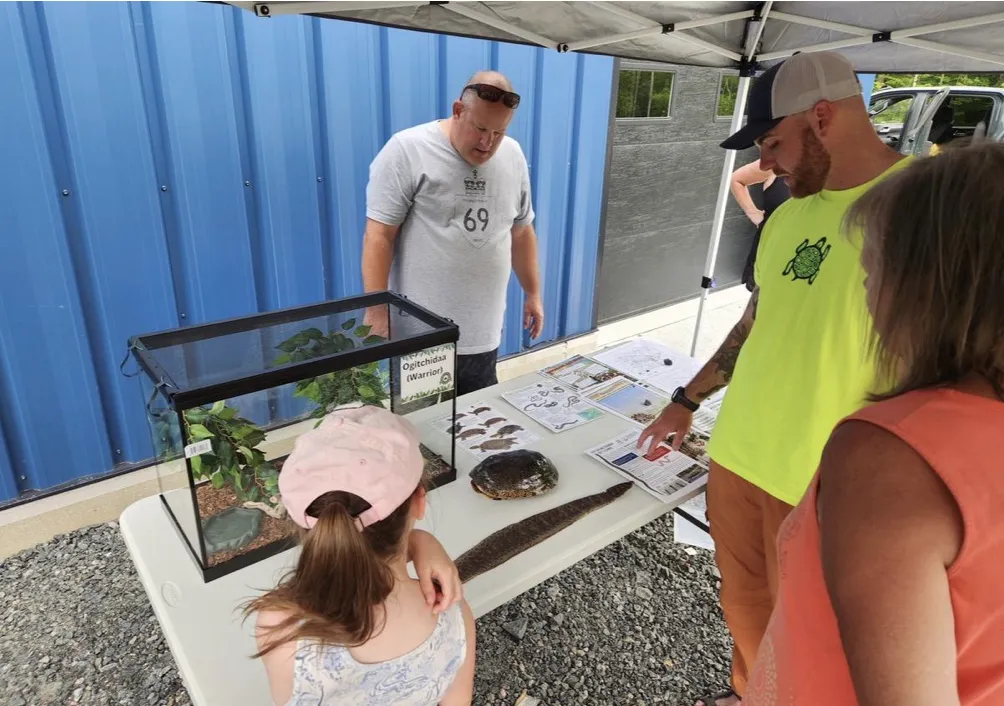 Aidan Maloney participating in a community outreach event under a tent, showcasing educational materials, a turtle shell, and a live reptile display case to interested community members.