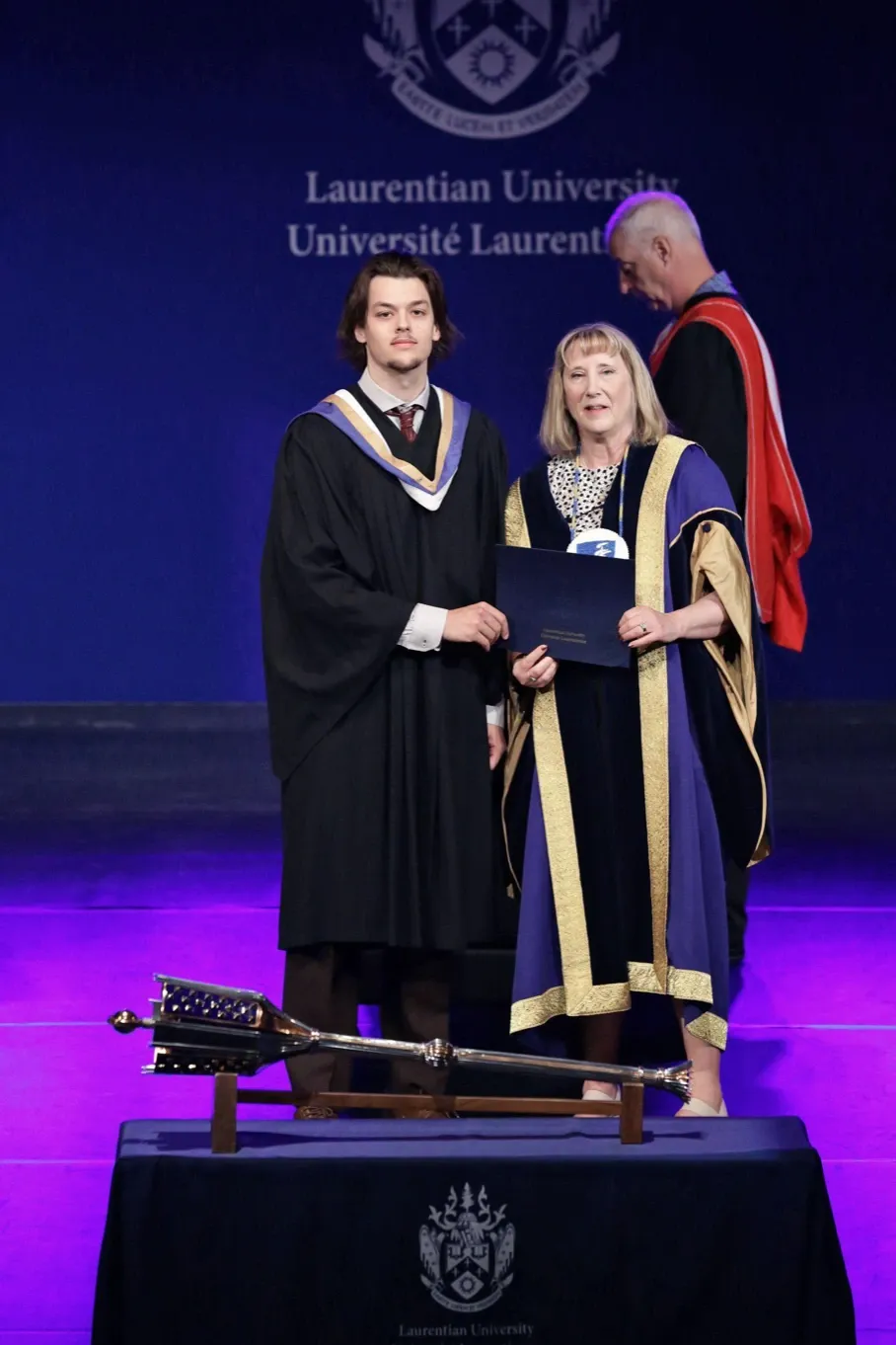 Laurentian University graduate Randy Kneer standing on stage in his black graduation gown and academic hood, smiling alongside Laurentian University President Dr. Lynn Wells, who is holding his degree certificate