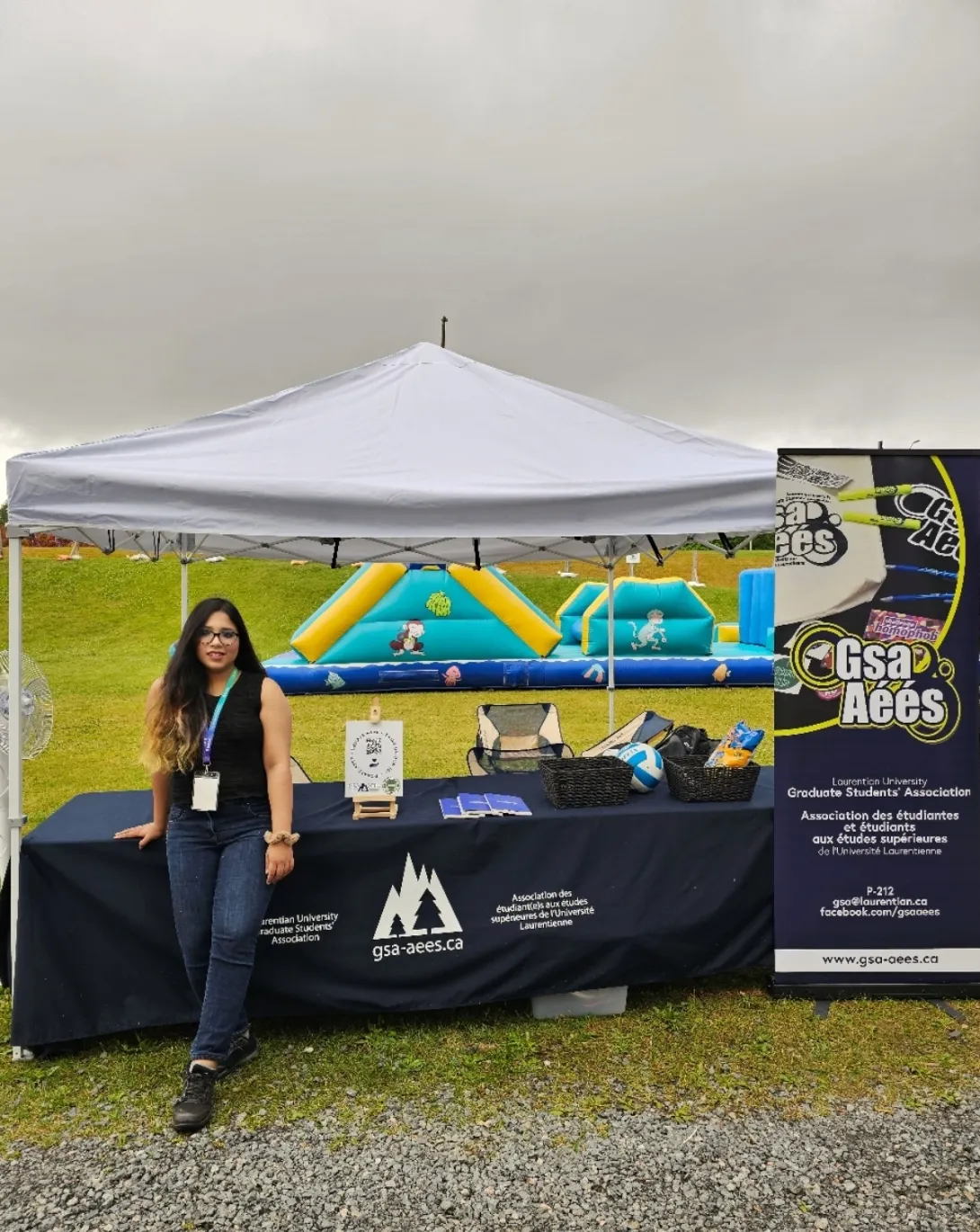 Samanta Saif Kristy posing at an outdoor Laurentian University Graduate Students' Association information table set up under a white pop-up tent.